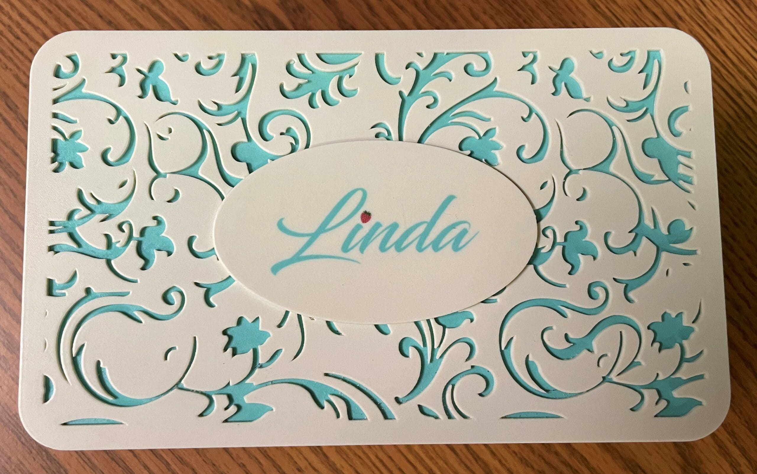 The personalized PolyJet jewelry-box lid features a recessed floral cut-out pattern in ivory, with a background layer of deep aqua, all printed as a single part. Forming the dot of the 