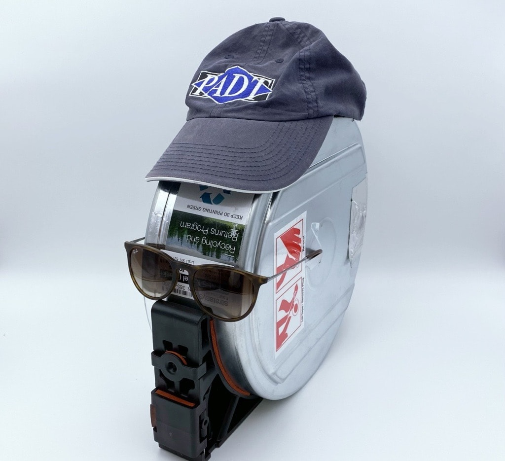 A canister of Stratasys 3D printing filament, with sunglasses and a PADT Inc. baseball cap on top.