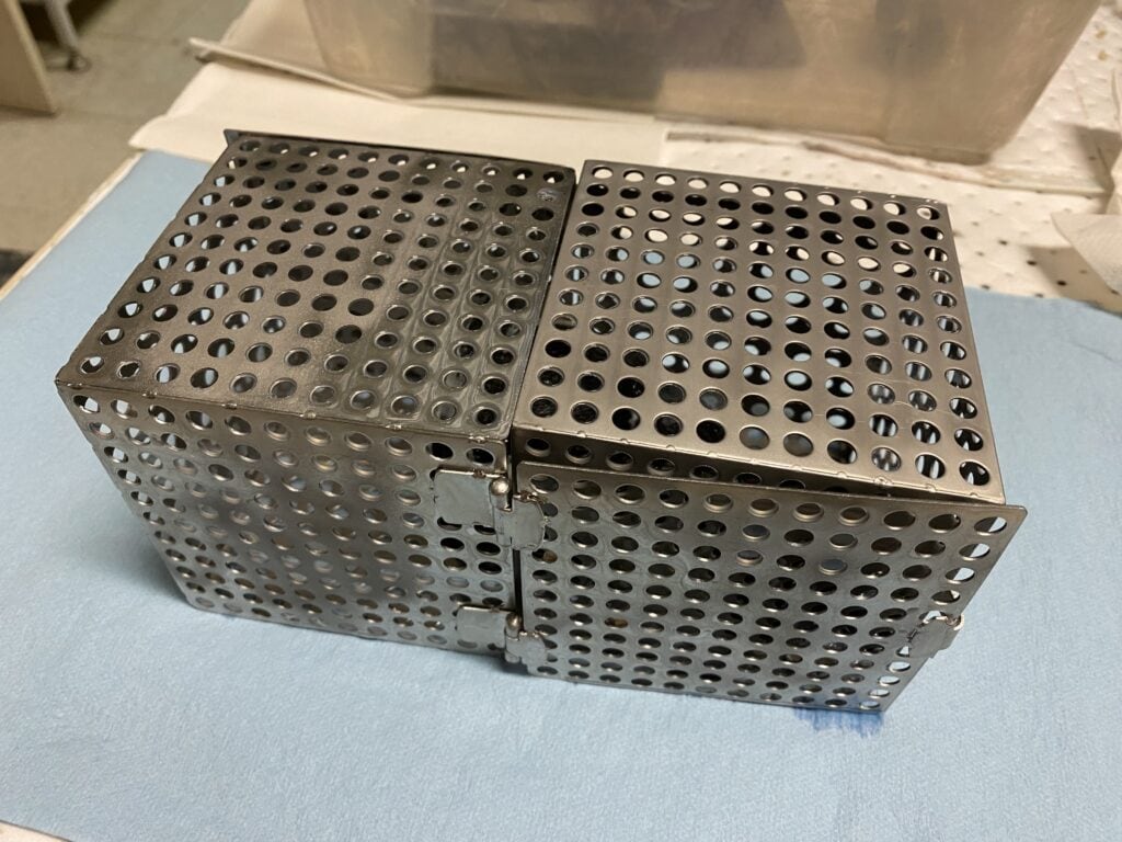 Two perforated small metal cube-shaped baskets, snapped together. They will hold a 3D printed part and keep the part from floating while support is being dissolved in an SCA tank.