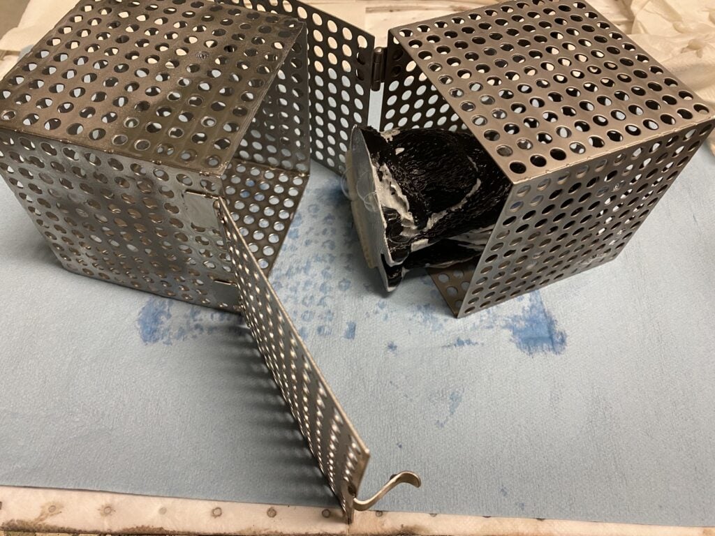 Two small perforated metal boxes, holding a 3D printed part, and almost snapped together. The box will stay on the bottom of an SCA tank while the printed part's support material is dissolved in sodium hydroxide.